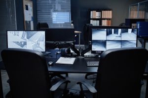 Empty security office showing two computer monitors displaying multiple surveillance camera feeds, office chairs positioned in front of desk, various documents and equipment arranged on tabletop