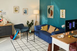 Modern living room interior featuring blue sofa, armchair, coffee table with cup and plate, computer monitor on desk, basketball and toy bat on floor, framed artwork on walls